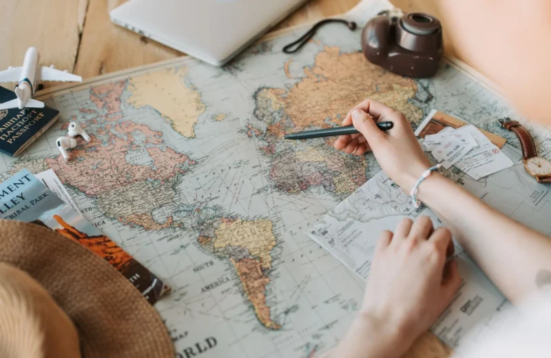 Hands marking destinations on a world map surrounded by travel essentials like passport and camera.