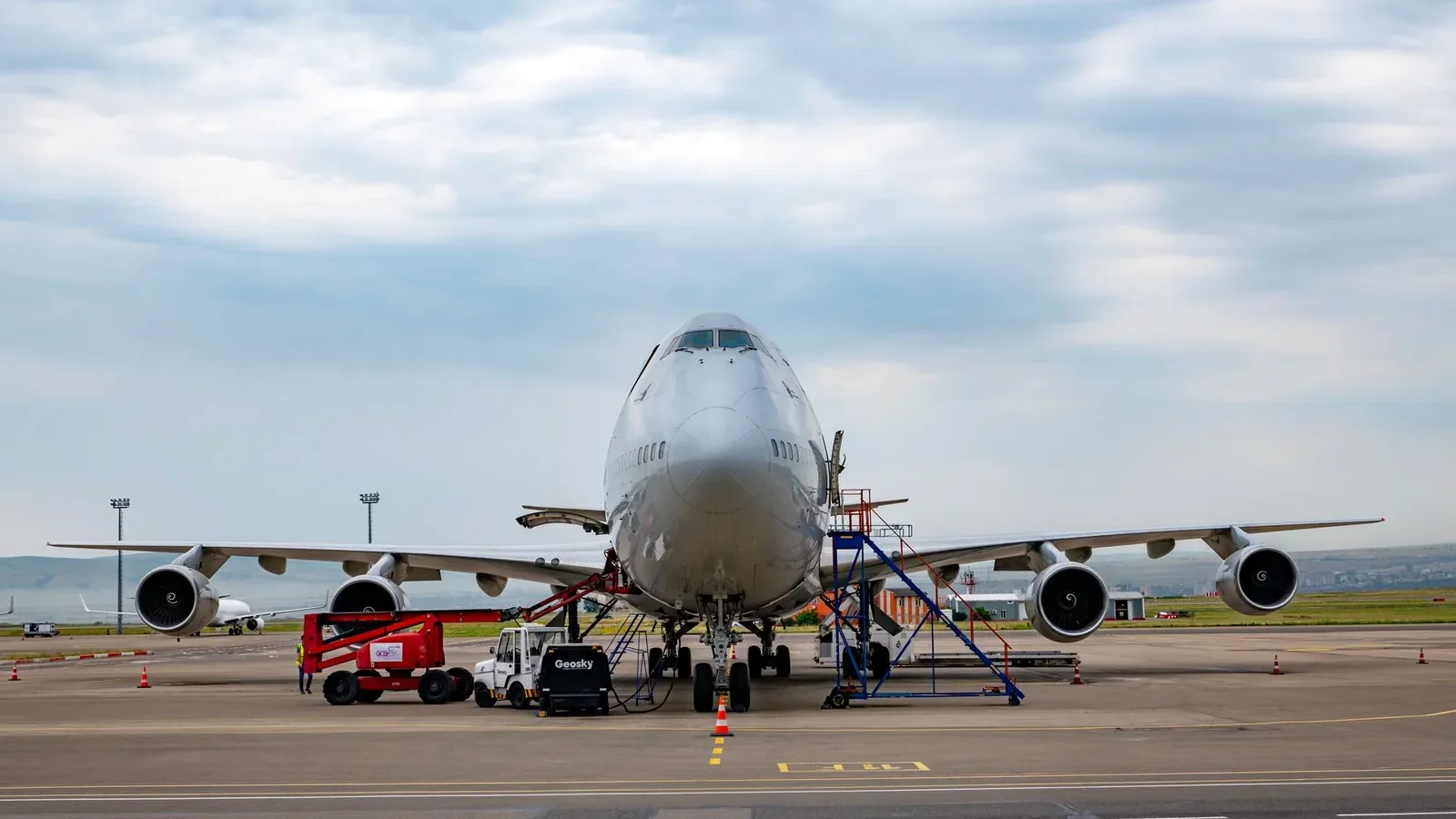 Front view of a Boeing 747 airplane at Tbilisi Airport with maintenance vehicles in the foreground.
