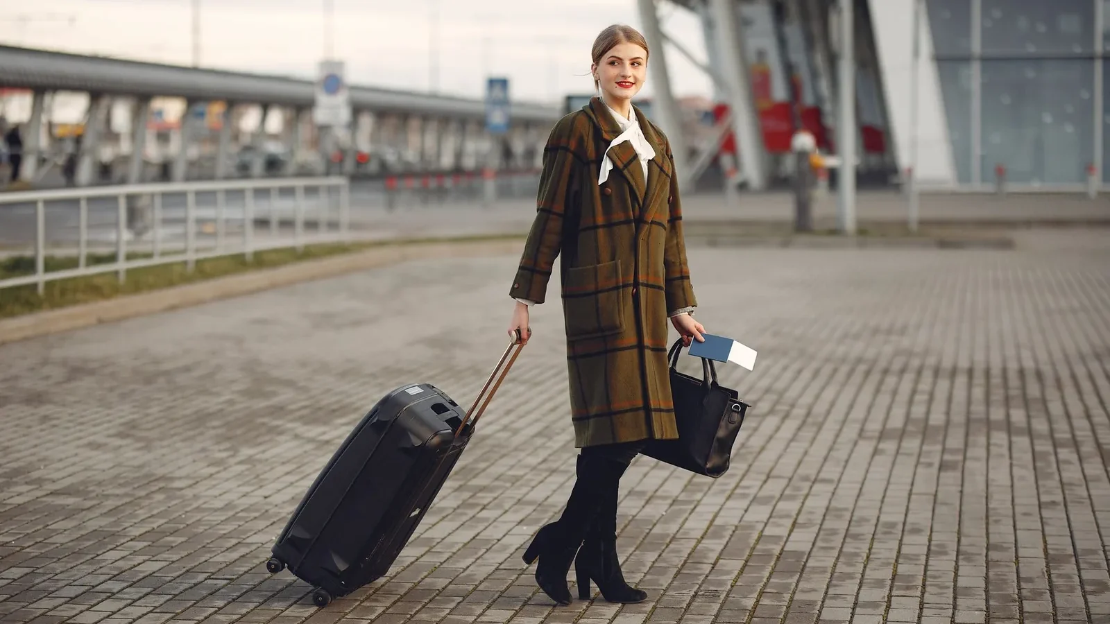 Full body young female passenger in trendy plaid coat with suitcase strolling on pavement near modern building of ariport terminal taking bag and documents.