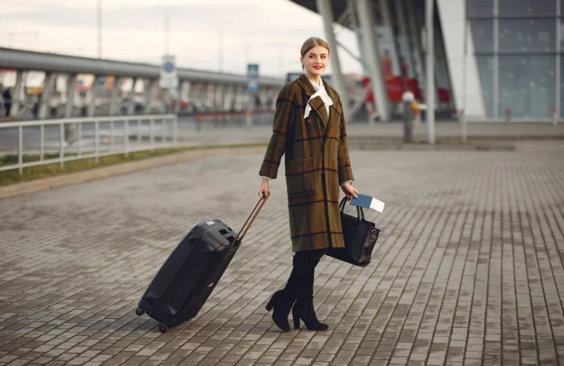 Full body young female passenger in trendy plaid coat with suitcase strolling on pavement near modern building of ariport terminal taking bag and documents.