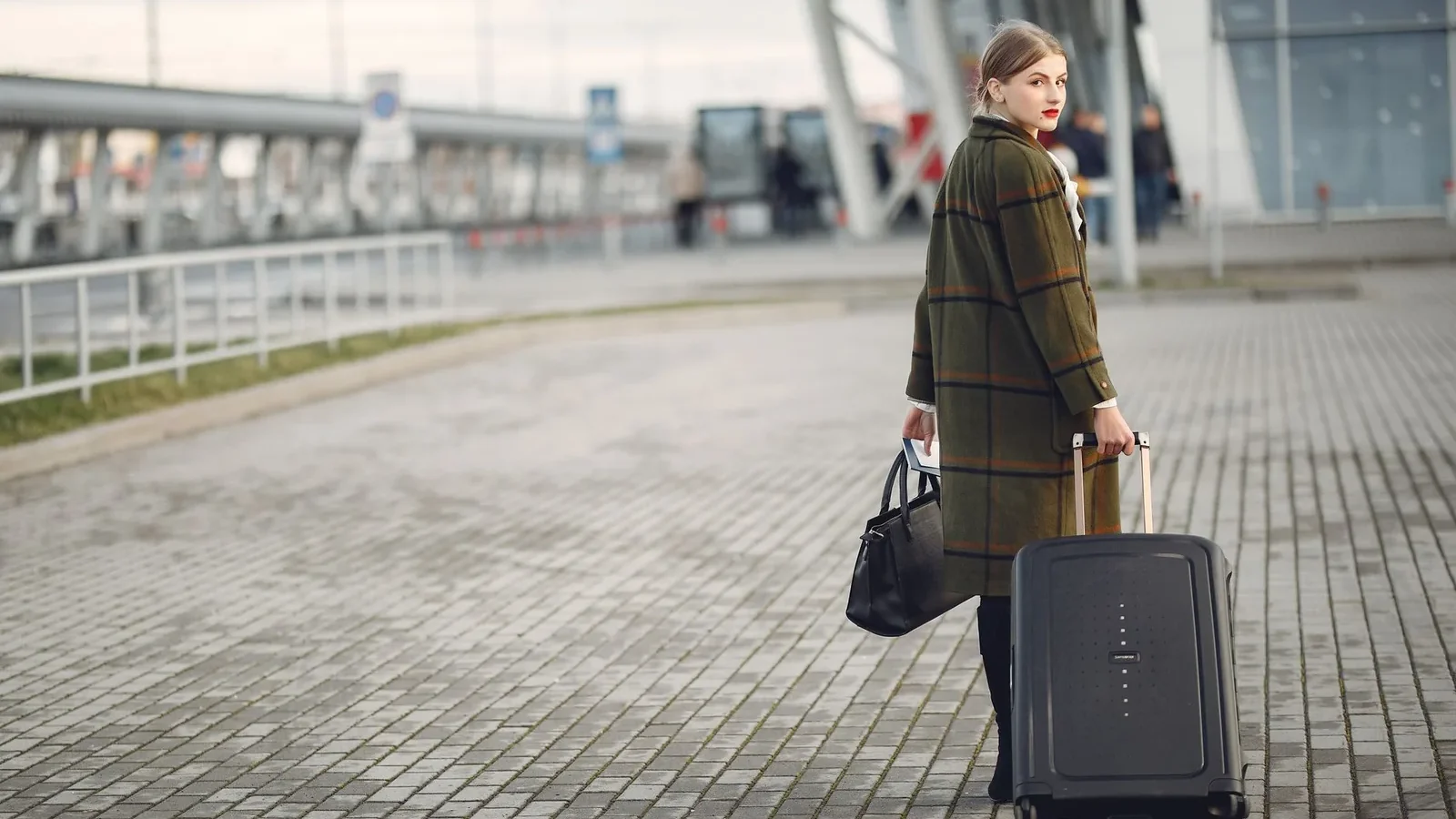 Full length of stylish female traveler in warm coat walking with suitcase and bag looking at camera after arrival against airport terminal.
