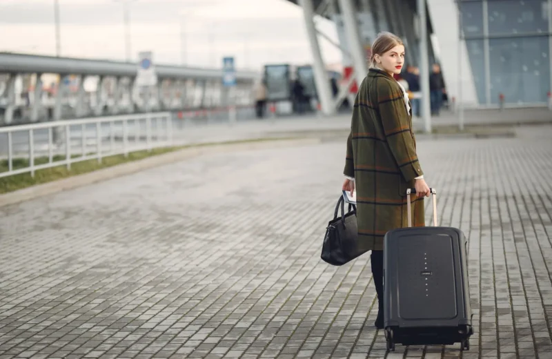 Full length of stylish female traveler in warm coat walking with suitcase and bag looking at camera after arrival against airport terminal.
