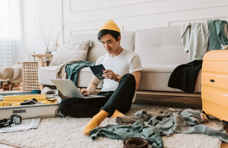 A young man in a messy living room preparing for travel with a laptop and passport.