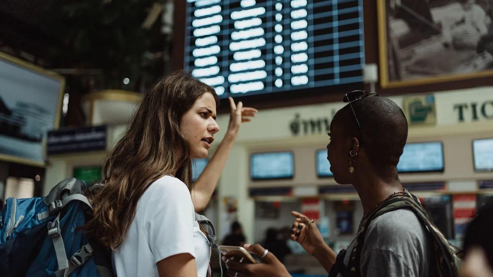 Two travelers with backpacks discuss in front of an airport timetable. Adventure awaits.