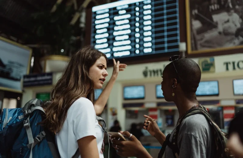 Two travelers with backpacks discuss in front of an airport timetable. Adventure awaits.