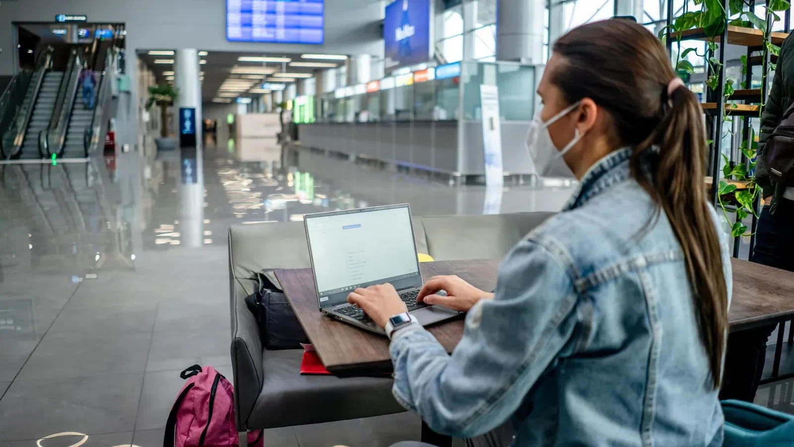 Side view of concentrated female freelancer in protective mask typing on netbook while sitting at table in modern airport terminal.