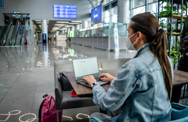 Side view of concentrated female freelancer in protective mask typing on netbook while sitting at table in modern airport terminal.