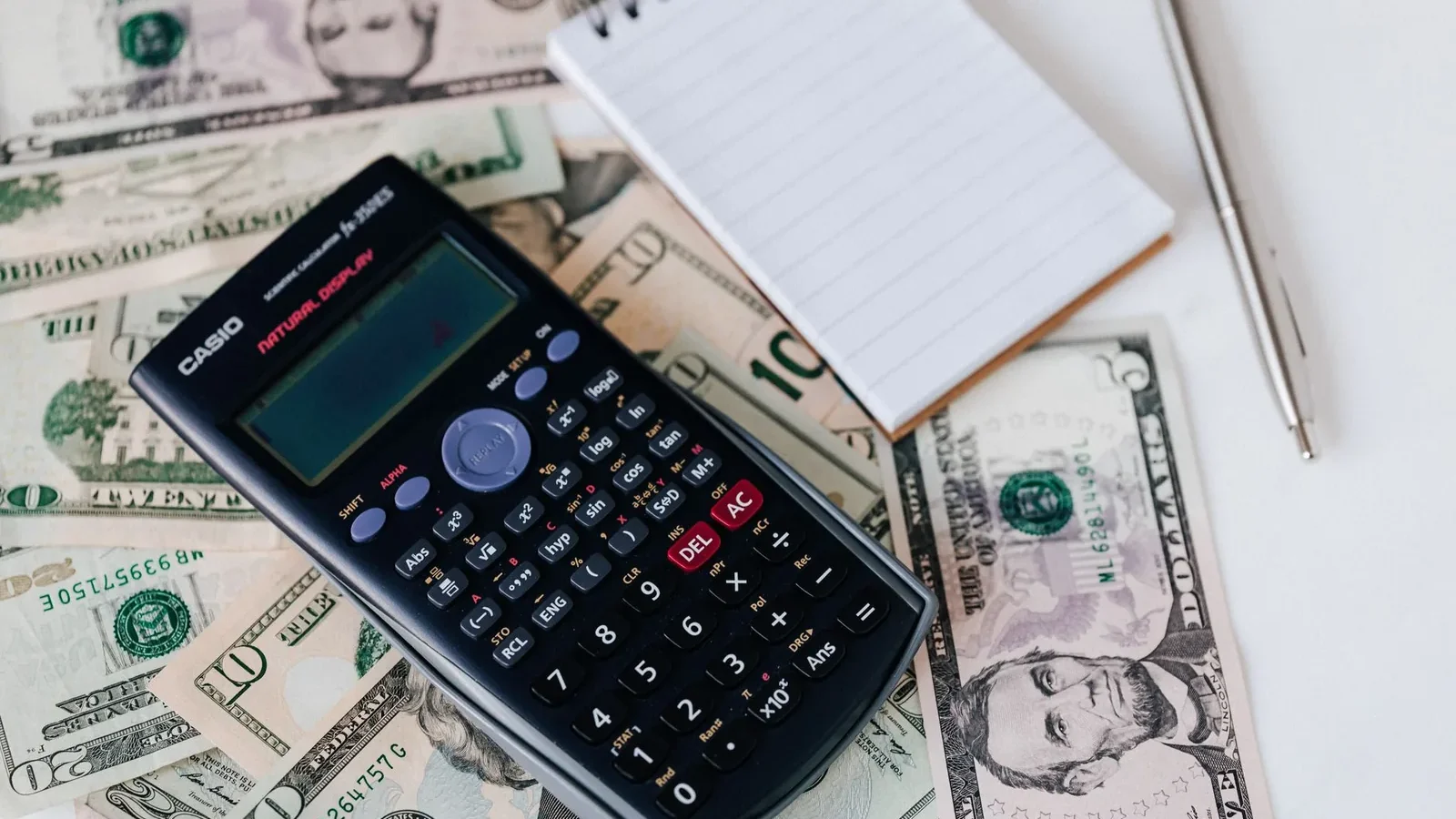 From above electronic calculator and notepad placed over United States dollar bills together with metallic pen for budget planning and calculation.