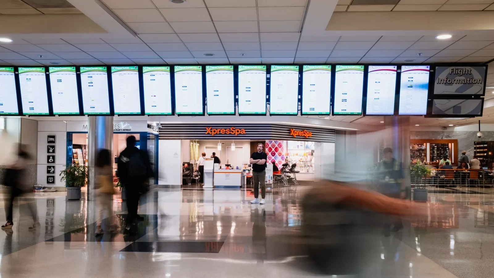 Busy travelers pass by an XpressSpa in Atlanta airport, under rows of flight information screens.