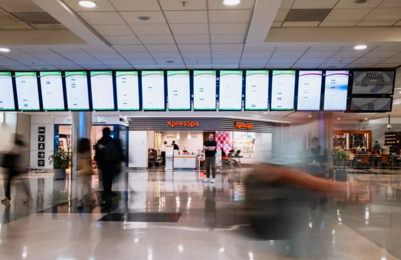Busy travelers pass by an XpressSpa in Atlanta airport, under rows of flight information screens.