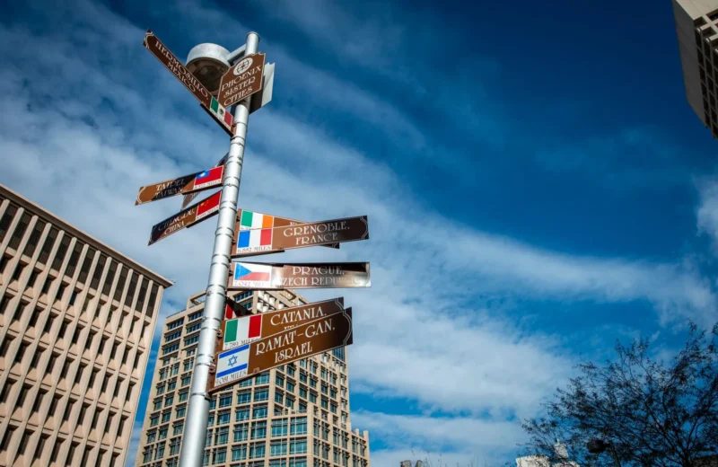 A low angle shot of a signpost featuring cities names and directions with office buildings in the background.