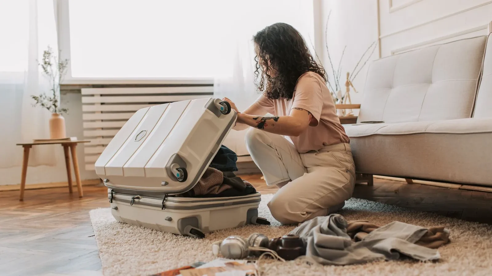 A woman packing her suitcase in a stylish living room, surrounded by clothes and travel accessories.