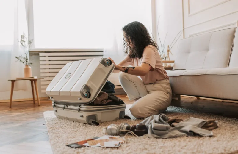 A woman packing her suitcase in a stylish living room, surrounded by clothes and travel accessories.