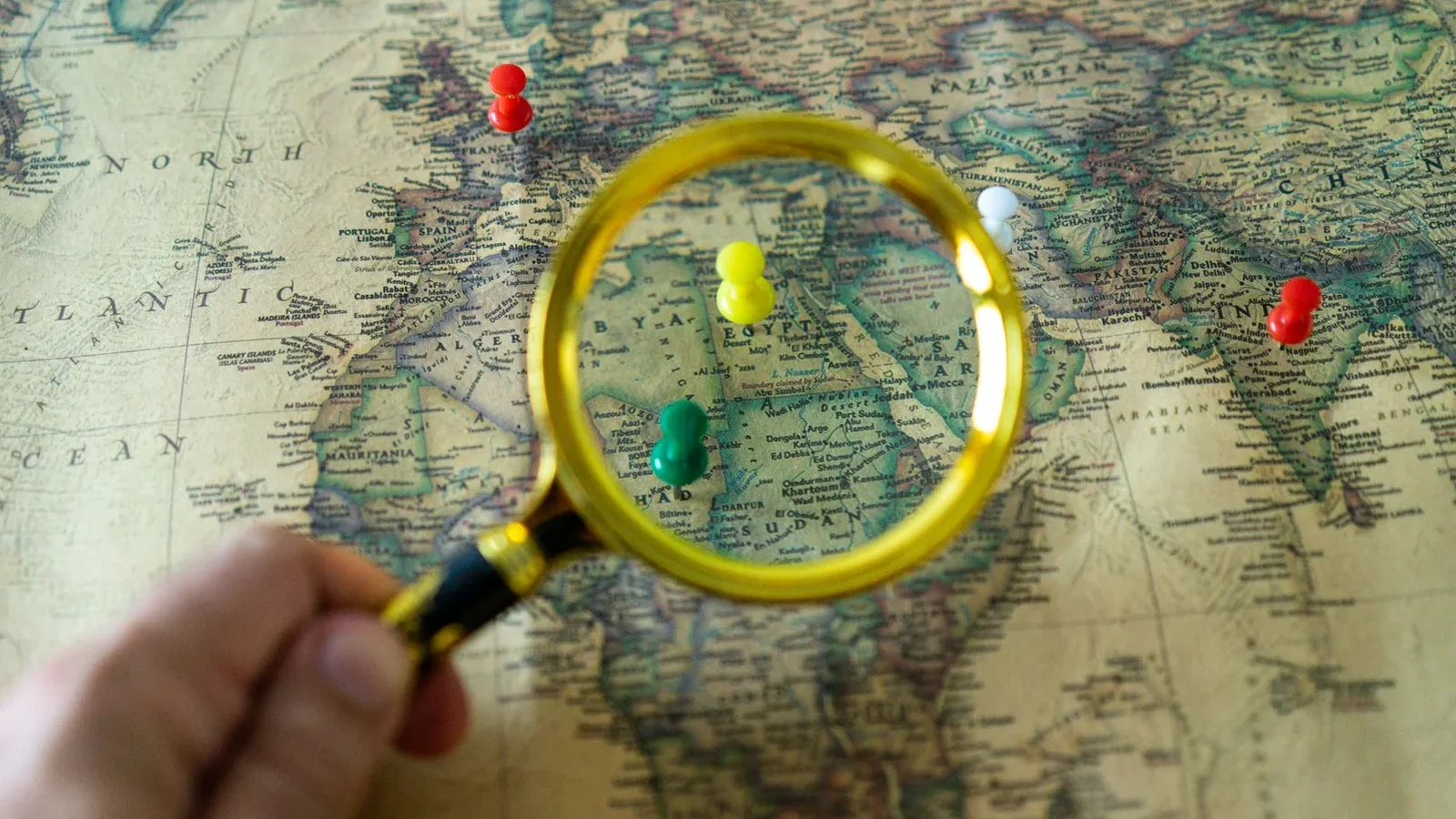Close-up of a hand holding a magnifying glass over a world map with colorful pins indicating travel destinations.