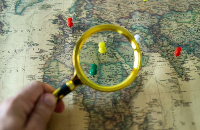 Close-up of a hand holding a magnifying glass over a world map with colorful pins indicating travel destinations.
