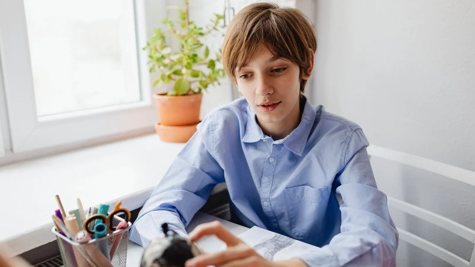 Teenager in blue shirt studying at a desk with a globe and stationery indoors near a window.