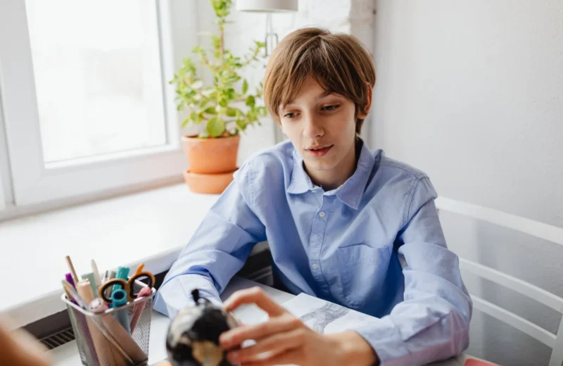 Teenager in blue shirt studying at a desk with a globe and stationery indoors near a window.
