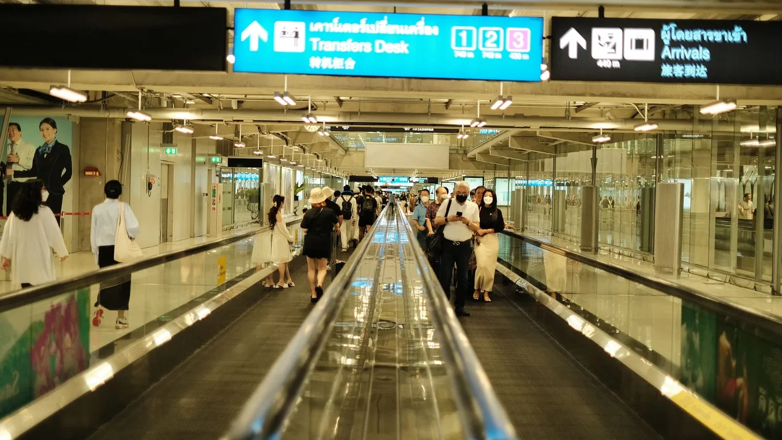 Passengers walk on a travelator in an airport arrival area, surrounded by signs and bright lighting.