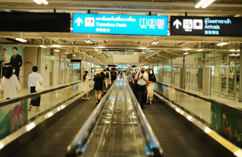 Passengers walk on a travelator in an airport arrival area, surrounded by signs and bright lighting.