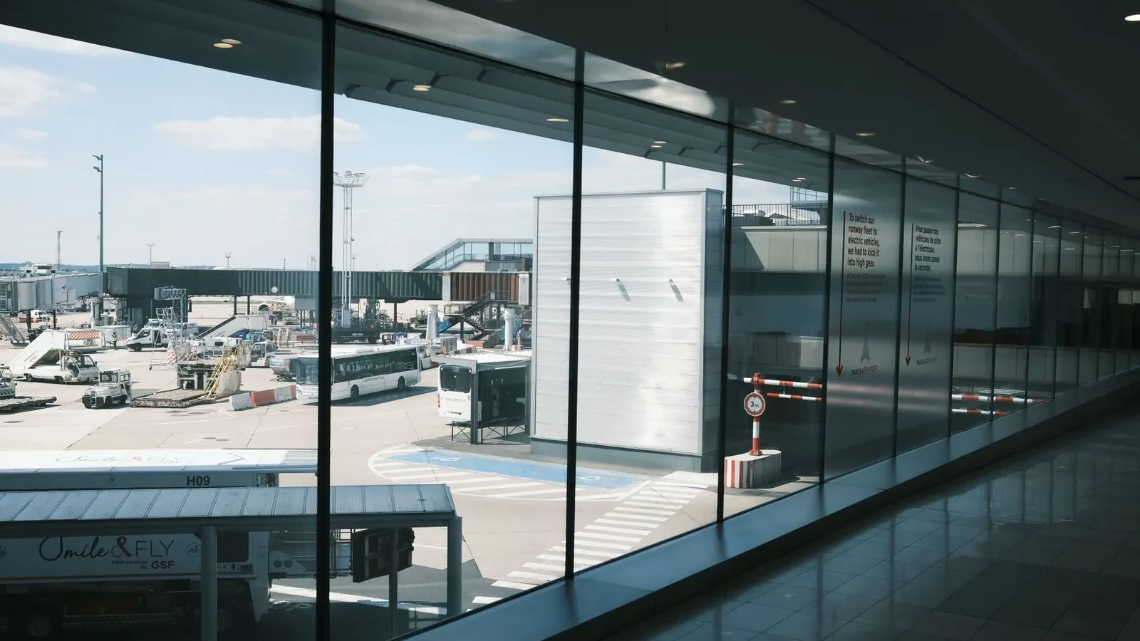 Wide view of an airport terminal hallway and tarmac, showcasing airplanes and buses.