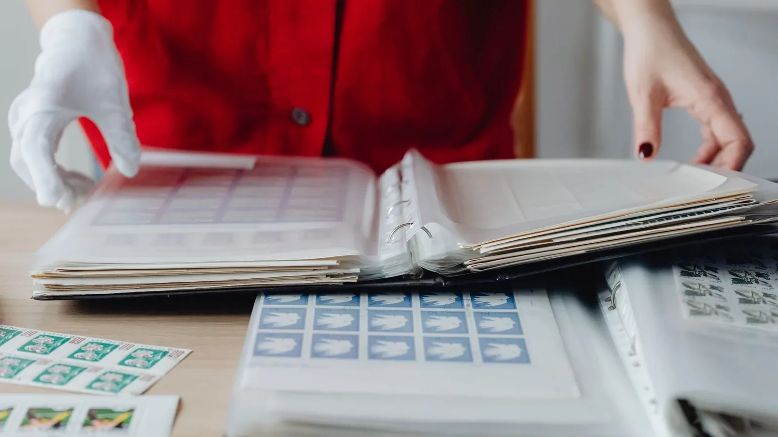 Person in red sorting a collection of vintage postage stamps using gloved hands on a wooden table.