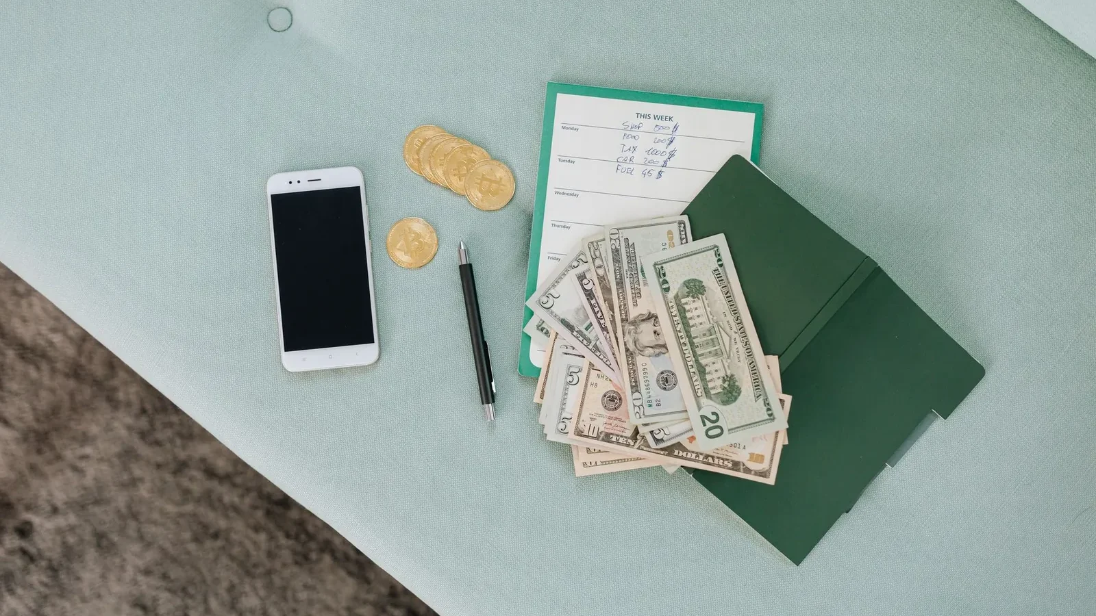Flat lay of a smartphone, US dollar bills, coins, and a planner on a green surface.