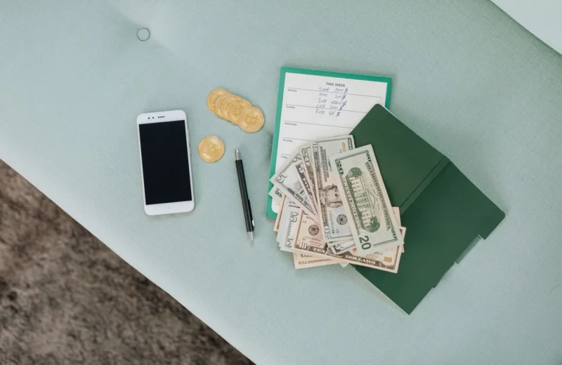 Flat lay of a smartphone, US dollar bills, coins, and a planner on a green surface.
