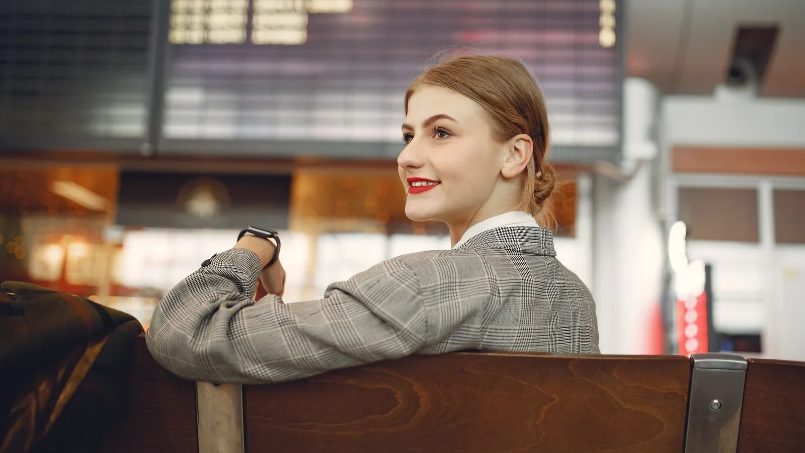 Optimistic young woman wearing jacket and smart watch and waiting for friend near departure board in waiting area of train station.