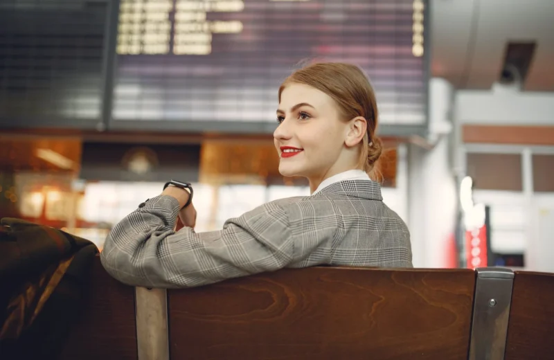 Optimistic young woman wearing jacket and smart watch and waiting for friend near departure board in waiting area of train station.