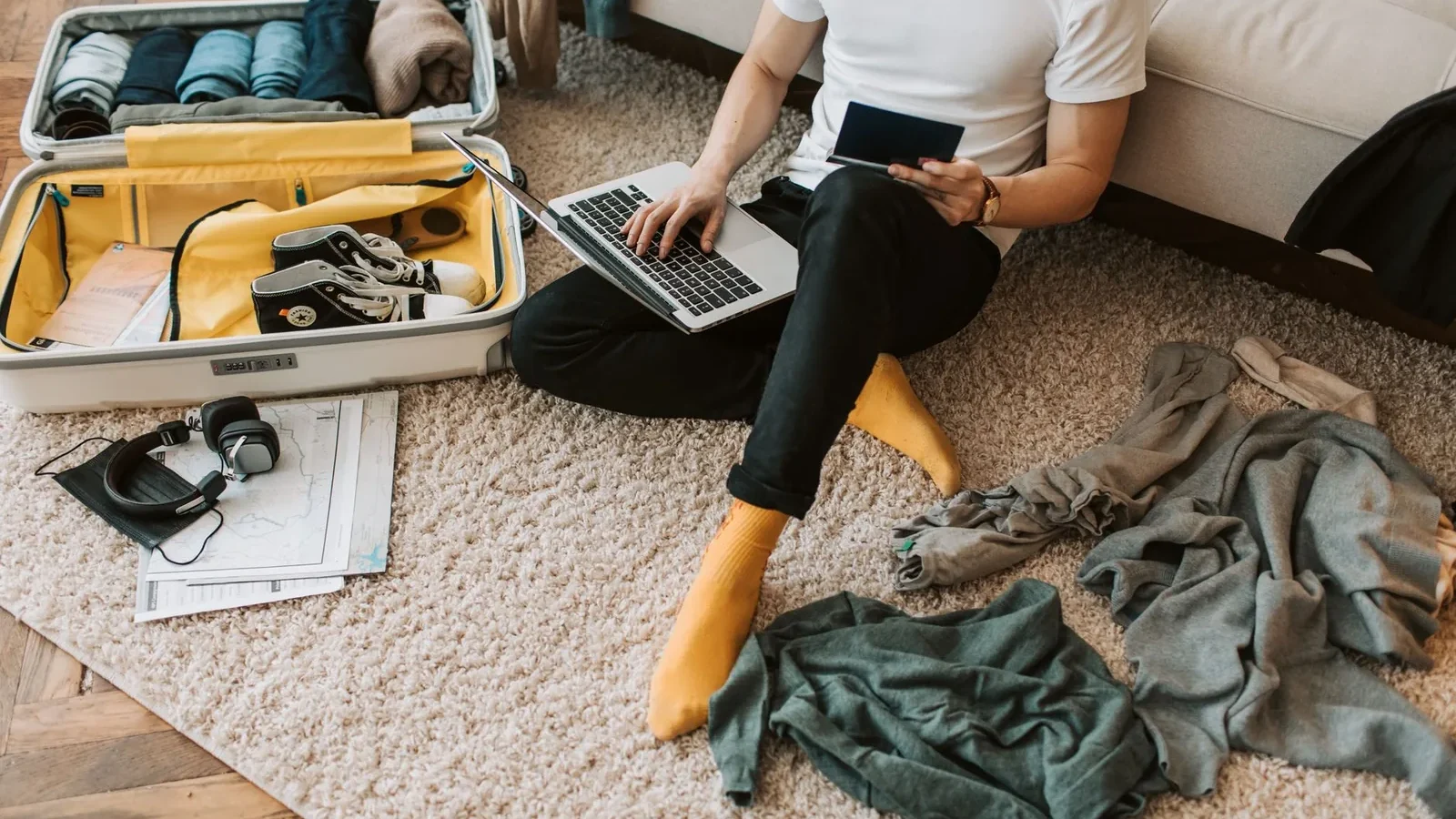 Man sits on floor packing suitcase while using a laptop, balancing travel and remote work.