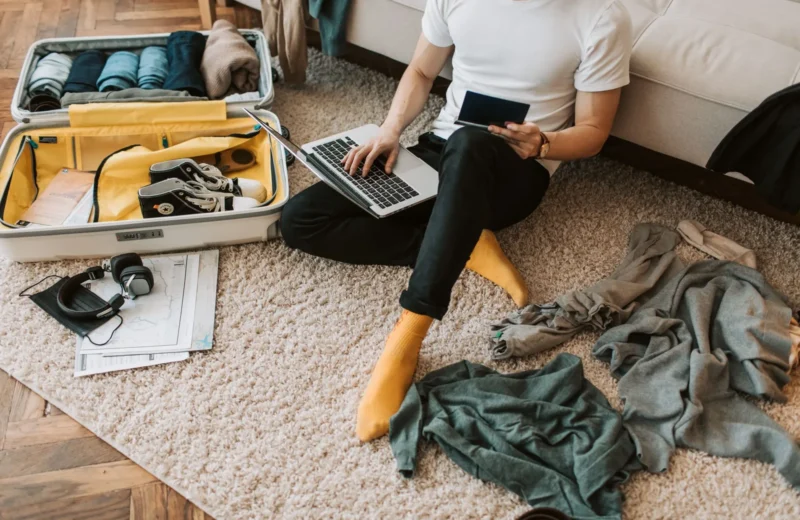 Man sits on floor packing suitcase while using a laptop, balancing travel and remote work.