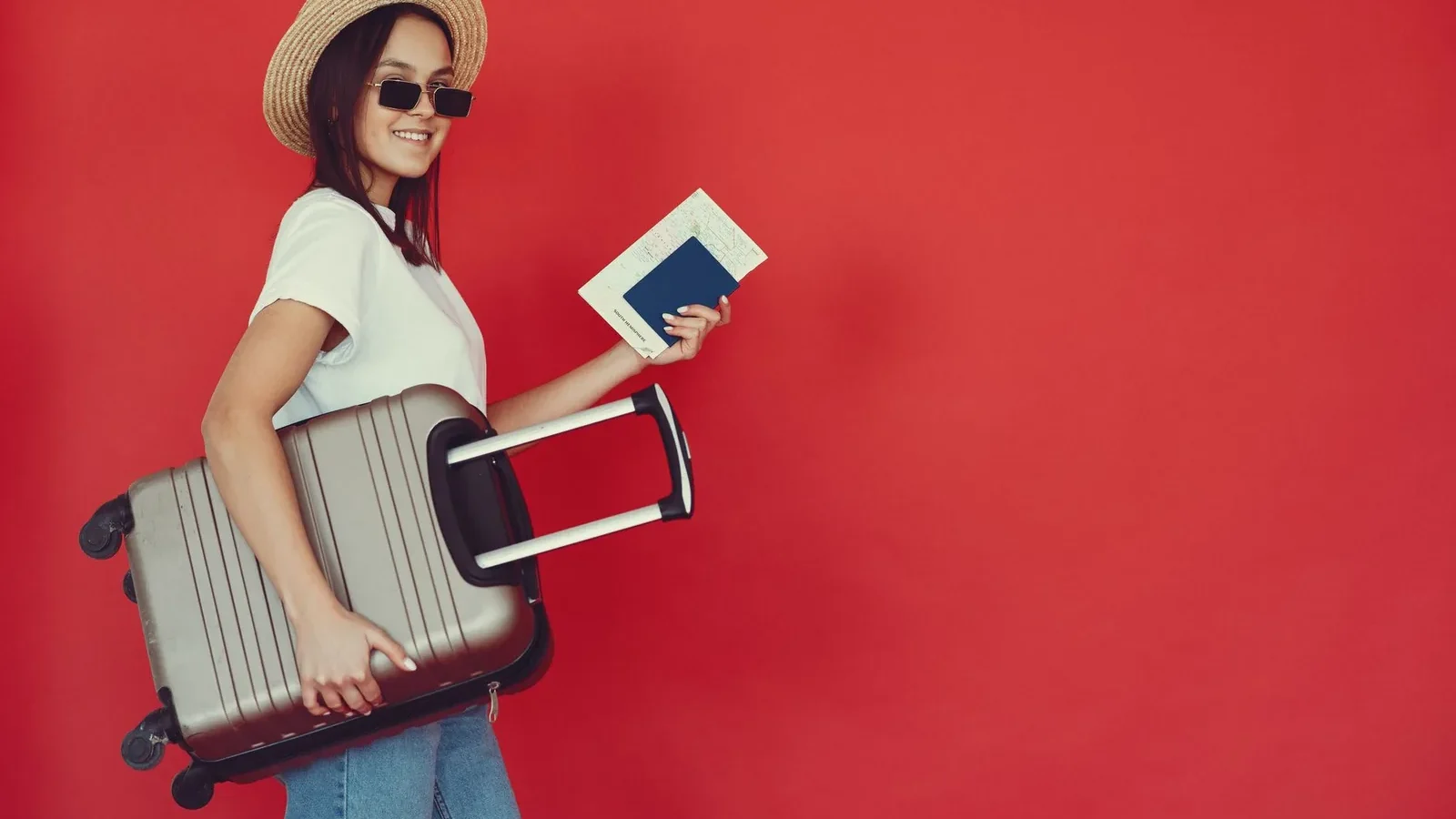 Side view of cheerful young woman with passport and tickets smiling and looking at camera while walking with baggage on red background.