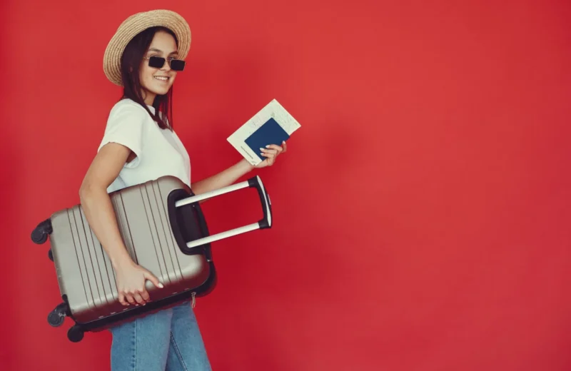 Side view of cheerful young woman with passport and tickets smiling and looking at camera while walking with baggage on red background.