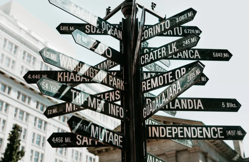 Close-up of a directional signpost with city names and distances in an urban setting.