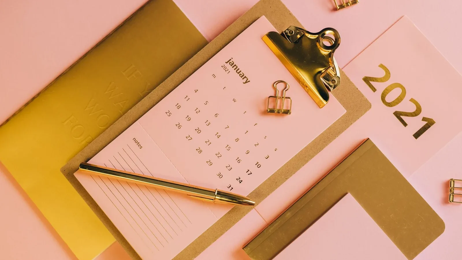 From above stylish workplace consisting of clipboard with calendar and golden notebook on pink background.