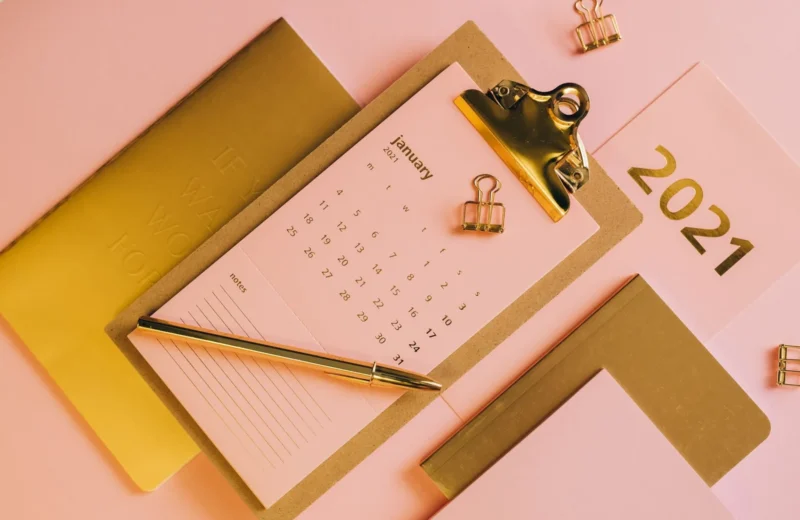 From above stylish workplace consisting of clipboard with calendar and golden notebook on pink background.