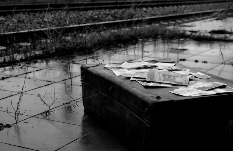 Black and white photo of an old suitcase and photos near train tracks, evoking nostalgia.