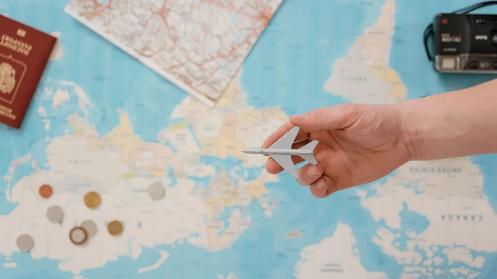 Hand holding a toy airplane over a world map, surrounded by coins, camera, and passport.