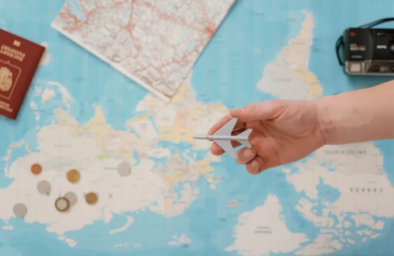 Hand holding a toy airplane over a world map, surrounded by coins, camera, and passport.