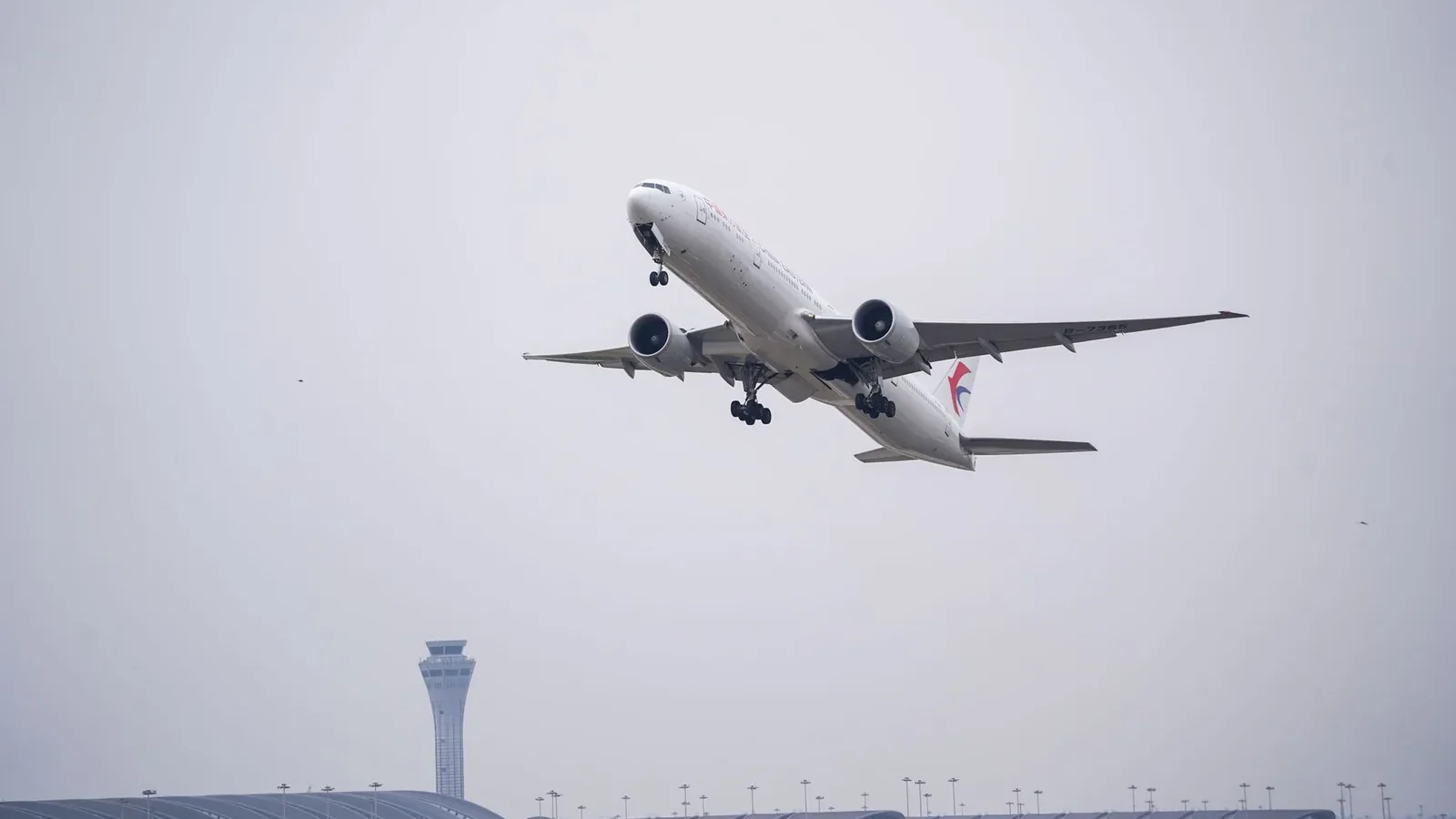 A commercial airplane ascends from an airport with a modern terminal and control tower visible.