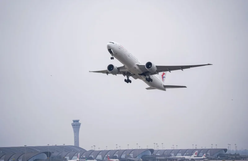 A commercial airplane ascends from an airport with a modern terminal and control tower visible.