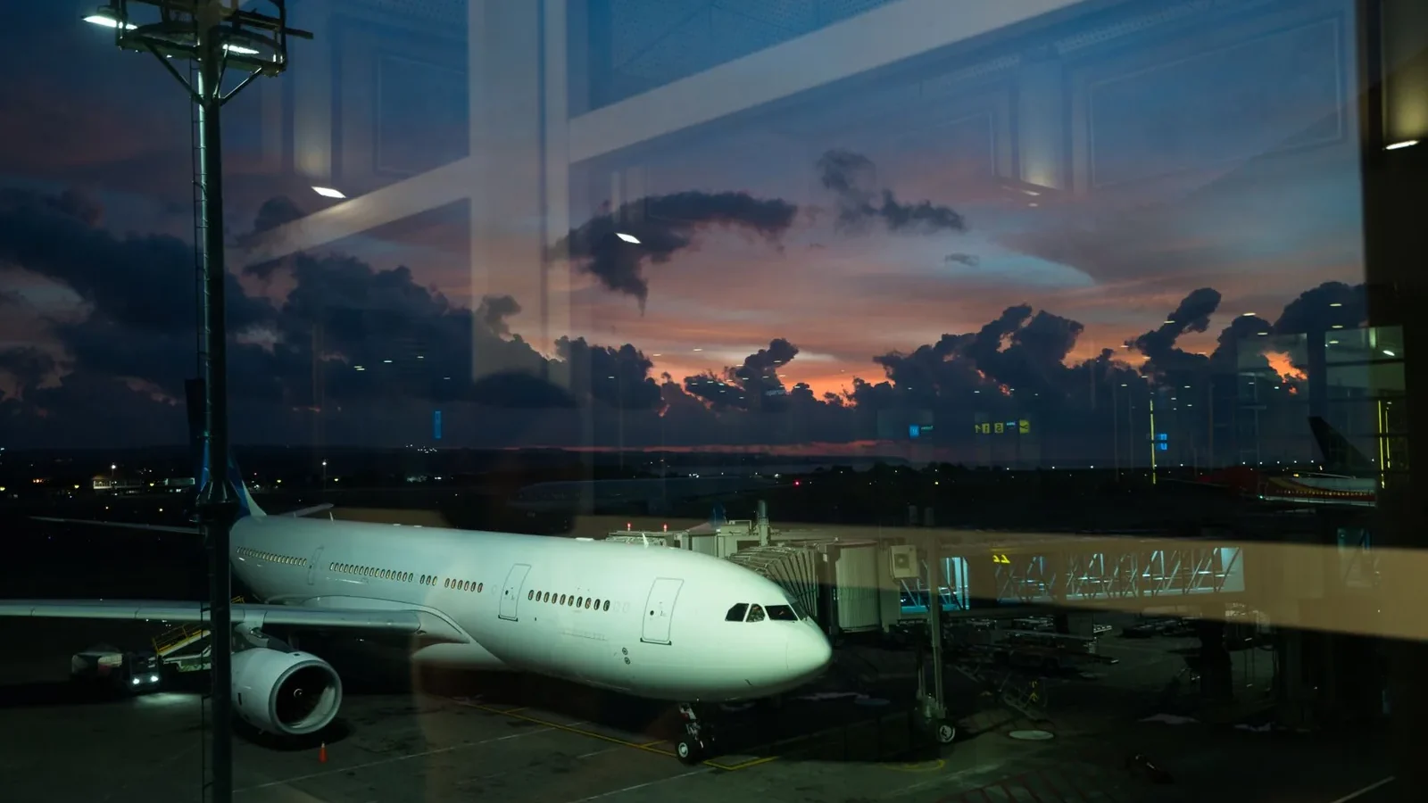 Through glass modern white plane connected to airport terminal with airbridge and ready for boarding against spectacular cloudy purple sky in late evening.