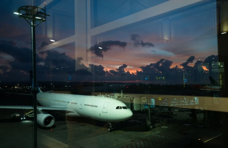 Through glass modern white plane connected to airport terminal with airbridge and ready for boarding against spectacular cloudy purple sky in late evening.