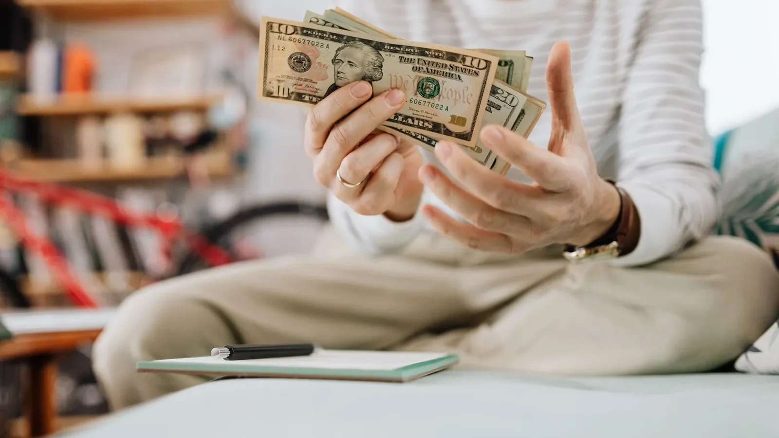 Man wearing casual clothes counts dollar bills while sitting on a sofa indoors.