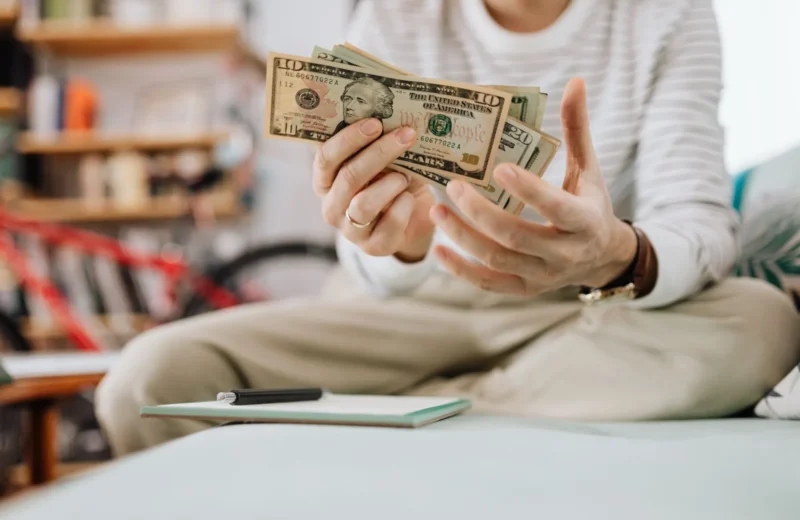 Man wearing casual clothes counts dollar bills while sitting on a sofa indoors.