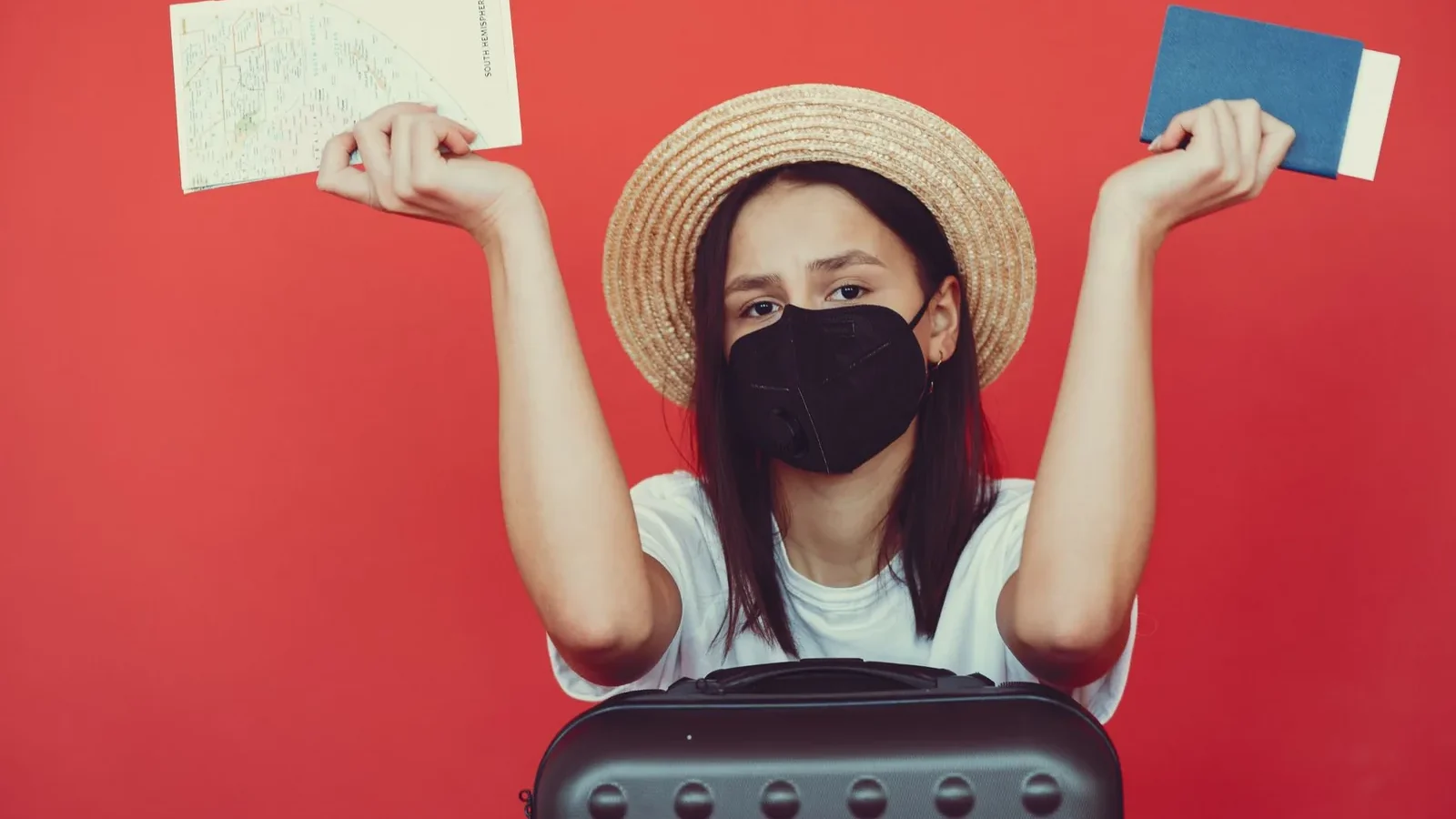 Upset young lady wearing straw hat and protective mask with map and passport leaning on black suitcase and looking at camera with disappointment on red background.