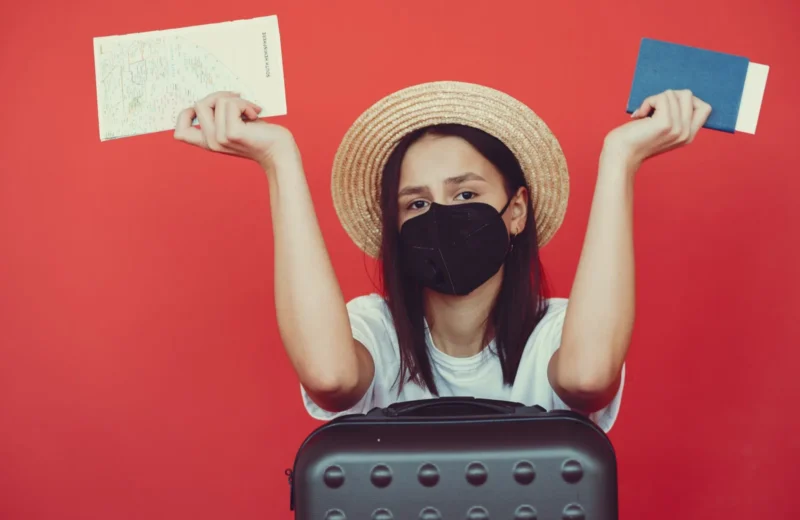 Upset young lady wearing straw hat and protective mask with map and passport leaning on black suitcase and looking at camera with disappointment on red background.