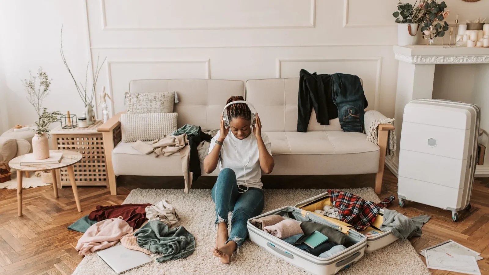 A woman packs a suitcase in her living room while listening to music, surrounded by clothes and a cozy atmosphere.