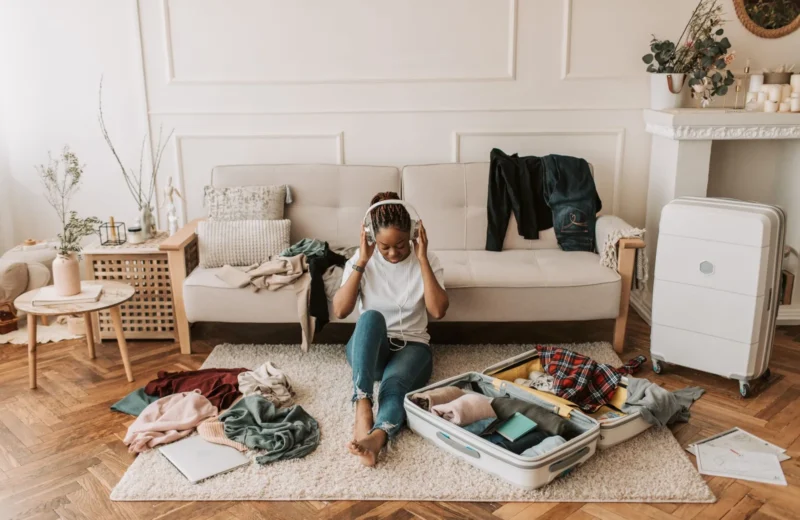 A woman packs a suitcase in her living room while listening to music, surrounded by clothes and a cozy atmosphere.