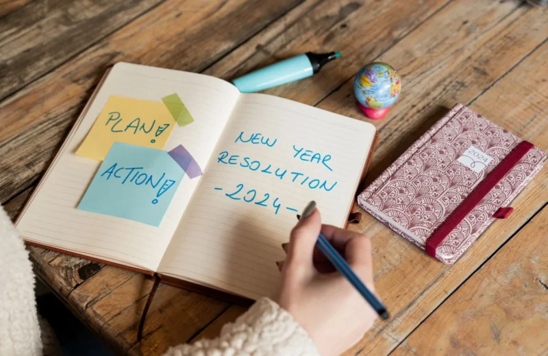 Person writing New Year 2024 resolutions in a notebook with sticky notes for planning and action.
